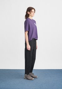 Fioletowa bawełniana koszulka z graficznym nadrukiem, w połączeniu z czarnymi, luźnymi spodniami i szarymi butami turystycznymi. Na neutralnym tle.