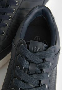 Sneakers blu navy con pelle liscia e dettagli perforati, lacci texturizzati e un logo sulla linguetta. Suola bianca.