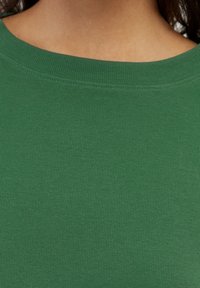Tessuto verde a coste con scollatura ampia, che mette in mostra un motivo a maglia testurizzato. Il materiale appare morbido e traspirante.