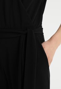 Gros plan sur un vêtement noir portefeuille avec une ceinture nouée et une main reposant dans la poche latérale.