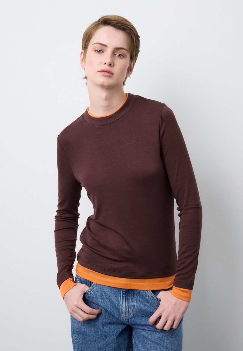 Junge Person mit kurzen Haaren, die ein braunes langärmeliges Hemd über einem orangen Oberteil und blauen Jeans trägt, steht vor einem weißen Hintergrund.