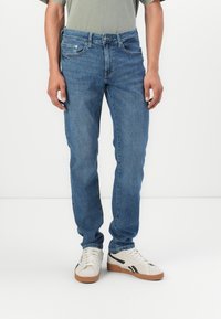 Jean en denim bleu avec une coupe droite, design à cinq poches et fermeture à bouton en métal. Portés avec des baskets blanches dotées d'une semelle en caoutchouc brun.