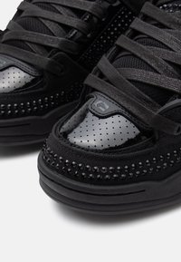 Des sneakers noires avec un dessus en tissu texturé, des accents en vernis noir brillant et des détails perforés, accompagnées de lacets plats noirs et de rivets.