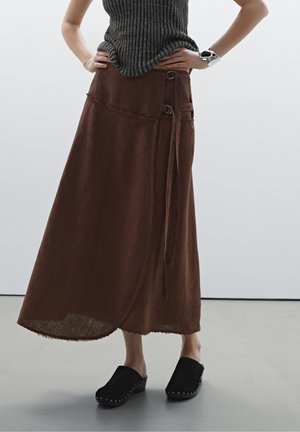 Person wearing a long brown wrap skirt with tie detail, black clogs with studs, and a sleeveless ribbed gray top with a silver cuff bracelet.