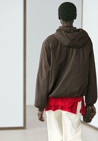 Mann in brauner Kapuzenjacke, rotem Lagenshirt und weißer Hose, der einen braunen Handschuh hält und mit dem Rücken zu einer schlichten hellen Wand steht.