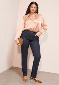 Sidenliknande beige skjorta med klassisk krage, kombinerad med mörka jeans. Modellen håller en liten brun kuvertväska och bär beige klackskor.