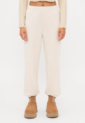 Kvinde iført cremefarvede bukser med elastisk talje, beige crop top og tan slip-on ankelstøvler mod en ensfarvet baggrund.