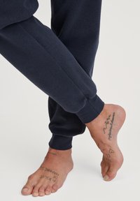 Pantalons de survêtement bleu marine avec des poignets côtelés, mettant en valeur un pied nu avec plusieurs tatouages. Le tissu a une texture douce et mate.