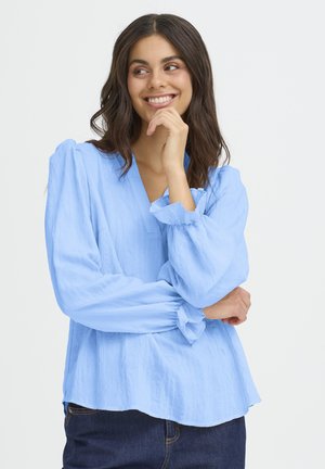 Junge Frau mit braunen Haaren, die eine hellblaue Langarmbluse und dunkle Jeans trägt, lächelt und schaut zur Seite, dabei hält sie die Hand nah an ihrem Gesicht.