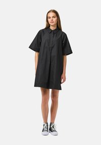Vestito camicia in cotone nero con colletto, maniche corte e vestibilità ampia. Indossato con sneaker alte in bianco e nero. Texture liscia.