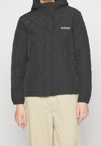 Person iført en sort quilted jakke med trykknapper, et logo på brystet og beige bukser, vist fra halsen til hoften.