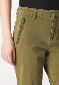 Olive spalvos kelnės su lygiu tekstūru, turinčios šoninį užtrauktuką, diržo kilpas ir klasikinį stilių, iš dalies parodytos su ranka.