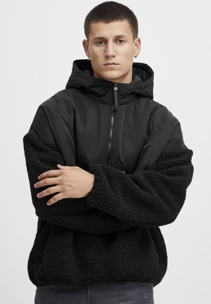 Zwarte hoodie met contrasterende pluizige fleece mouwen en een gladde romp. Heeft een rits aan de voorkant en een comfortabele pasvorm.