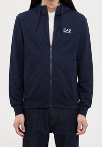 Marinblå hoodie med dragkedja i mjukt tyg, med framdragkedja, sidfickor, justerbar huva och broderad EA7-logotyp på bröstet.