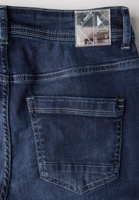 Dark denim jeans featuring a single back pocket, concealed stitching, and a shiny label with geometric design near the waistband.