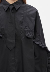Robe chemise noire avec col pointu, cravate noire étroite et détails à volants sur la poitrine et les manches.