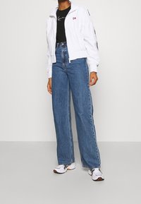 Bílá bomber bunda na zip s elastickými manžetami, nošená přes černé tričko. Džínsy s vysokým pasem a širokými nohavicemi modré barvy a bílé tenisky s detaily.