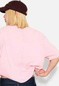 Světle růžové oversized tričko s krátkým rukávem a kulatým výstřihem, doplněné o tmavě hnědou kšiltovku. Z bavlněného materiálu, ležérní design.