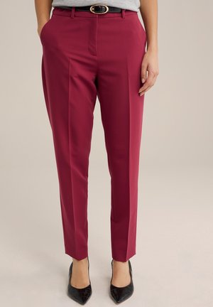 Vrouw draagt op maat gemaakte bordeauxrode broek met voorplooien, zwarte riem met gouden gesp, grijze top en zwarte pumps met spitse neus.