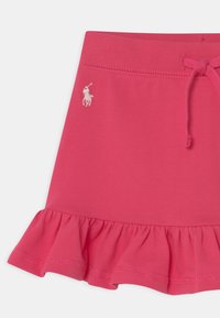 Rosa bomullskjol med volangkant, med texturerad yta och en vit broderad logga på sidan. Justerbar dragsko i midjan.