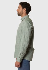 Camisa de manga larga verde claro hecha de tela texturizada, con cuello abotonado y detalles en los puños, usada con jeans azules.