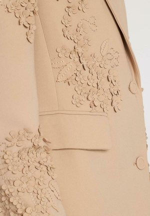 Blazer beige su misura con dettagli in applicazione floreale sulle maniche e sulla parte anteriore, forma strutturata, con chiusura a un bottone e tasca applicata.