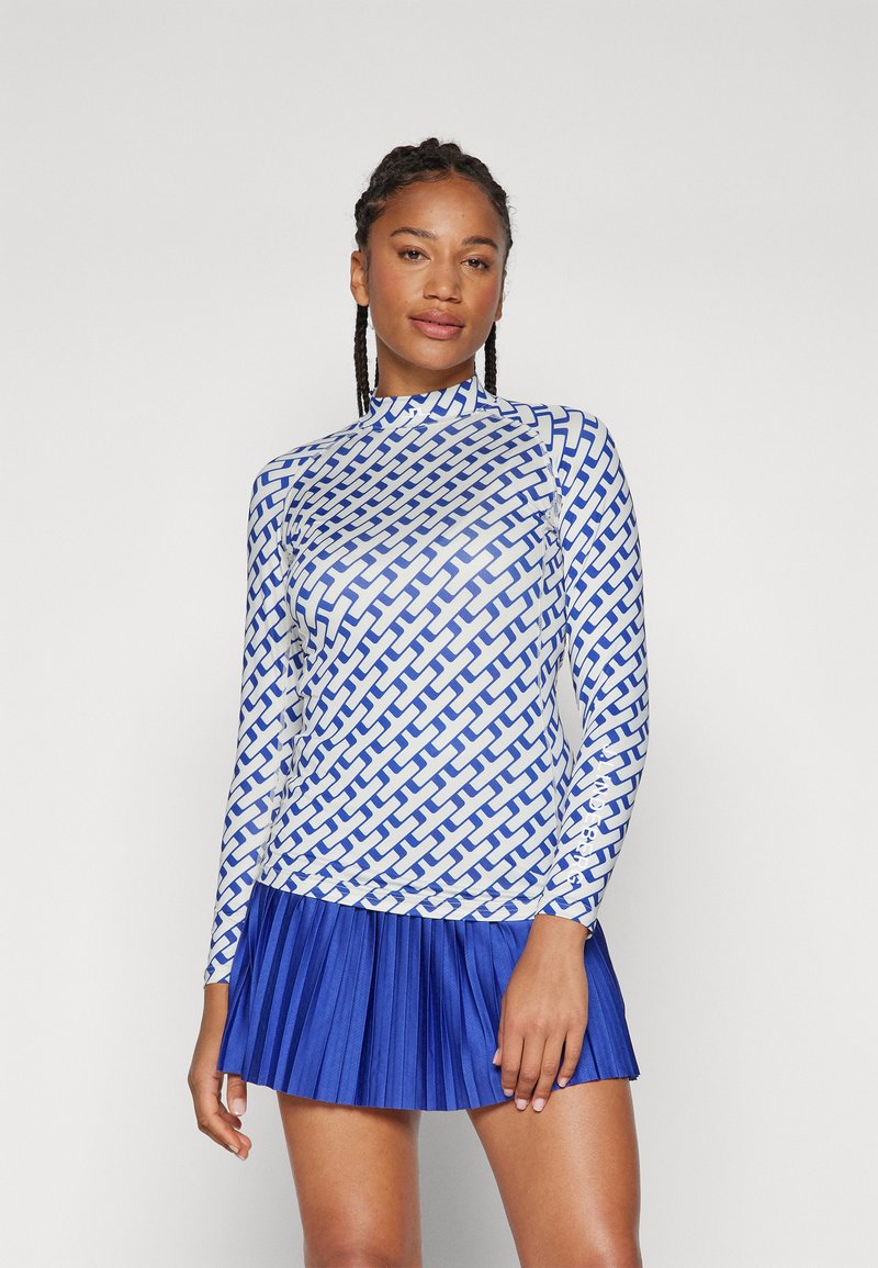 Long-sleeve shirt in white print with blue geometric pattern, paired with a pleated blue skirt. Both feature smooth, stretchy fabric.