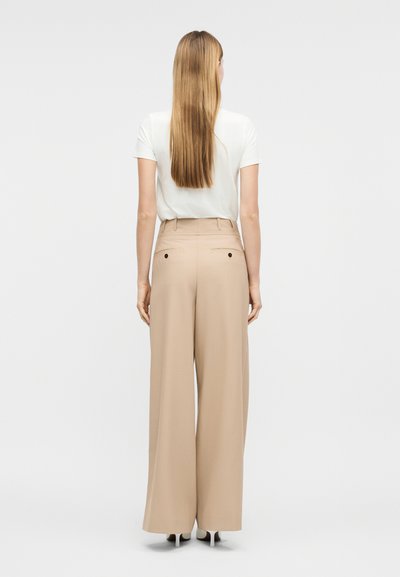 Γυναίκα με μακριά ξανθά μαλλιά στέκεται με την πλάτη στην κάμερα, φοράει λευκή μπλούζα με κοντά μανίκια, μπεζ παντελόνι φαρδύ στο κάτω μέρος και λευκά παπούτσια με ψηλό τακούνι.