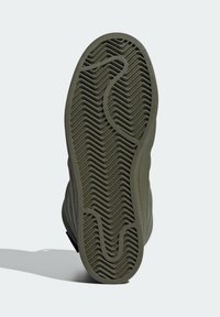 Sole olivově zeleného tenisky s chevronovým vzorem a zakřivenými drážkami, vrhající stín na světlém pozadí.