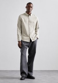 Ljust beige knapp-up skjorta med en bröstficka, i kombination med vida grå byxor och svarta skor. Slät tyg i en avslappnad passform.