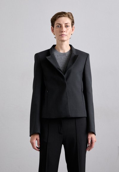 Sort skræddersyet blazer med en struktureret silhuet, skarpe revers og vinklede skuldre, båret over en grå sweater, parret med matchende bukser.