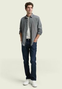 Camisa de botones gris sobre una camiseta blanca, combinada con jeans azul oscuro y zapatillas blancas. El modelo posa con las manos en los bolsillos.