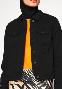 Veste en denim noire avec fermeture à boutons, deux poches poitrine et un col classique. Associée à un pull côtelé orange et une jupe à motifs.