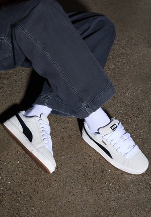 Person iført mørke bukser, hvide sokker og hvide Puma-sneakers med sort logo, som sidder på en ru betonoverflade.