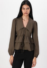 Blouse vert olive avec un décolleté en V profond, un design à volants en couches et des liens à la taille. Dotée de manches longues bouffantes et de poignets froncés.