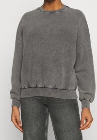 Persona che indossa un felpa oversized grigia lavata con polsini e orlo a costine, abbinata a jeans di denim grigio scuro.