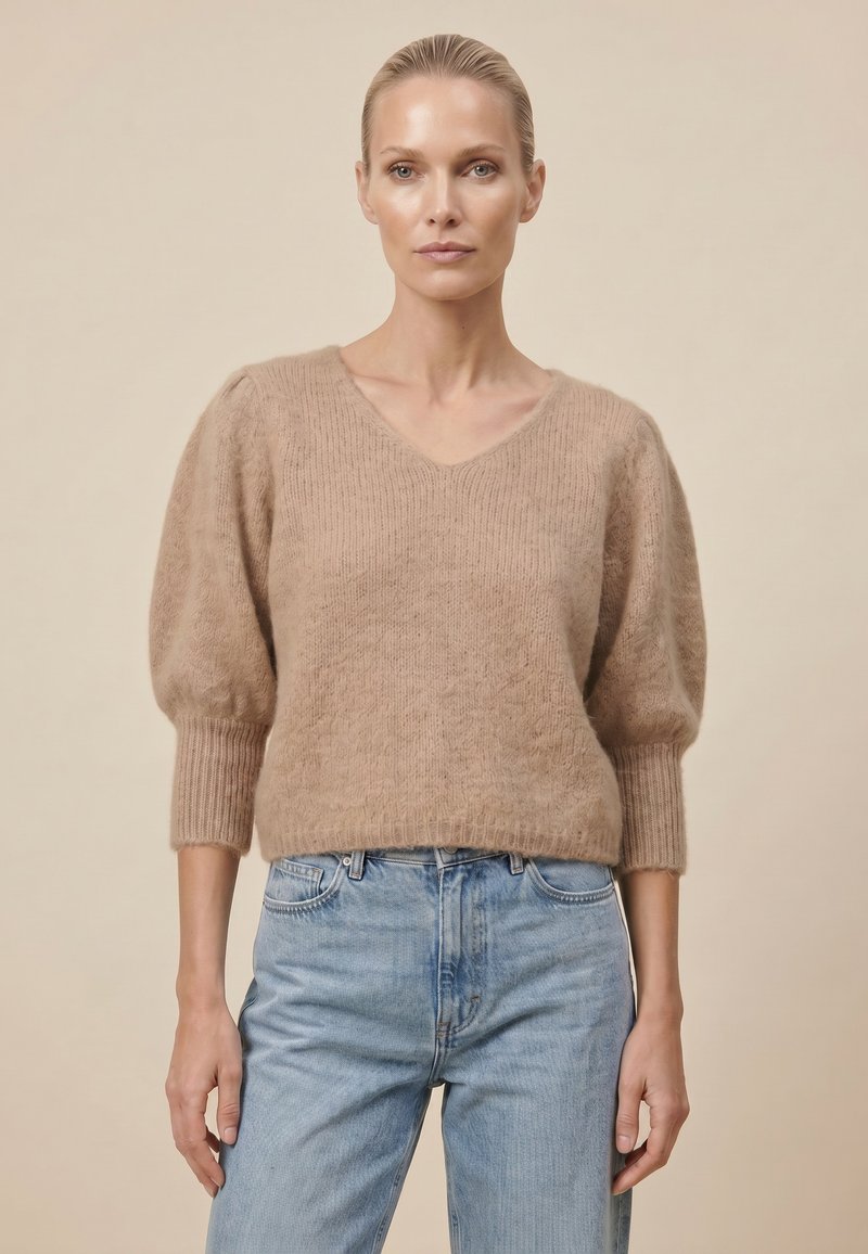 Femme aux cheveux slickés en arrière portant un pull beige duveteux avec des manches ballons et un jean bleu clair, debout devant un fond uni.