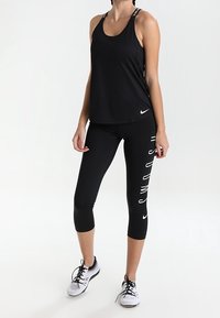 Top sportivo nero con spalline sottili e logo Nike; abbinato a leggings Capri neri con grafica bianca "SWOOSH". Scarpe da corsa grigie.