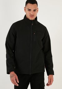 Chaqueta softshell negra con cuello alto, cremallera frontal y bolsillo en el pecho con cremallera. Incluye bolsillos laterales con cremallera y un diseño de marca minimalista.