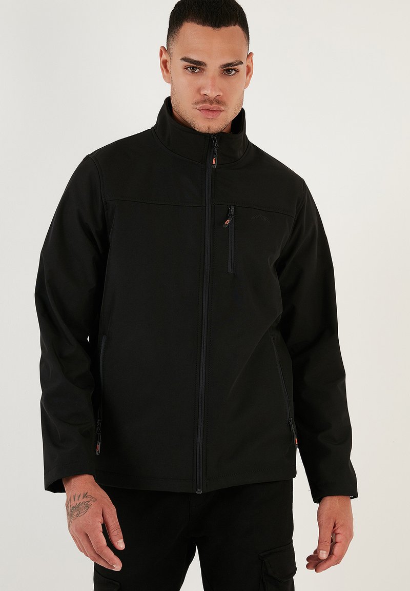 Chaqueta softshell negra con cuello alto, cremallera frontal y bolsillo en el pecho con cremallera. Incluye bolsillos laterales con cremallera y un diseño de marca minimalista.
