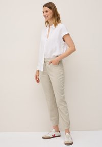 En kvinna med vågigt hår ler medan hon står, klädd i en vit t-shirt med kort ärm, beige byxor och vita sneakers, framför en neutral bakgrund.
