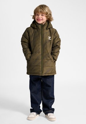 Jong kind met blond haar, draagt een olijfgroene jas met capuchon, donkerblauwe spijkerbroek en beige sneakers, glimlachend met handen in de zakken.