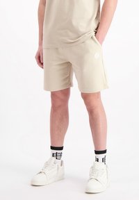 Beige shorts van zachte stof, met zijzakken en een logo patch. Draagt met witte sneakers en sokken met zwarte accenten.