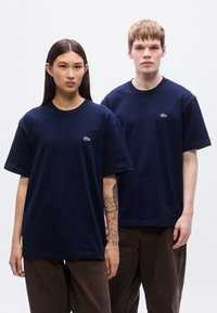 Μπλε βαμβακερά μπλουζάκια με κοντά μανίκια και λαιμόκοψη, με μικρό πράσινο λογότυπο κροκόδειλου στο αριστερό στήθος. Ορατό τατουάζ του μοντέλου.