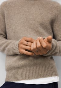 Beżowy sweter z dzianiny z prążkowanym dołem i mankietami. Miękka tekstura i dopasowany krój. Ręce z widocznymi tatuażami chwytają się nawzajem.