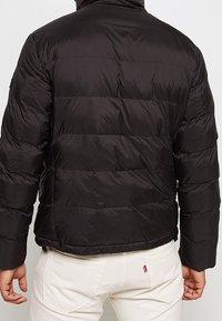 Person som bär en svart vadderad jacka och off-white byxor sedd bakifrån, stående mot en enkel ljus bakgrund.