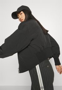 Black padded bomber jacket with wide sleeves, ribbed cuffs, and hem. Worn with pinstriped pants, showcasing a sleek, minimalist design.