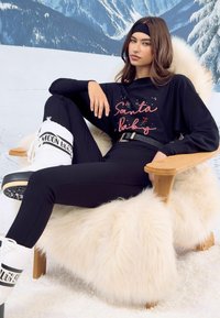 Fekete hosszúujjú póló "Santa baby" flitterekkel, fekete leggingsek, fehér hócsizmák fekete díszítéssel, egy bolyhos széken ül.