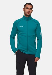 Teal fleecejack met een hoge kraag, ritsluiting en twee zijzakken met rits. Heeft contrasterende stiksels en een logo op de borst.