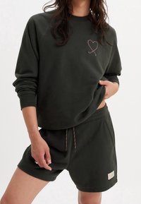 Tmavě zelená mikina s dekorativním srdcovým designem a odpovídajícími šortkami. Materiál je jemný, s duhovými stahovacími šňůrkami a malou nášivkou s logem.